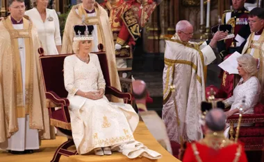Camilla wearing a crown during a formal coronation ceremony, seated with clergy in ornate robes.