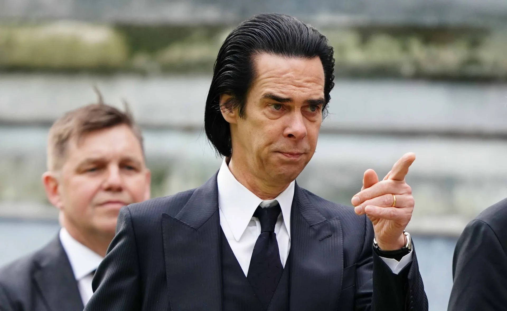 A man in a black suit and tie gestures with his hand, with another person blurred in the background.