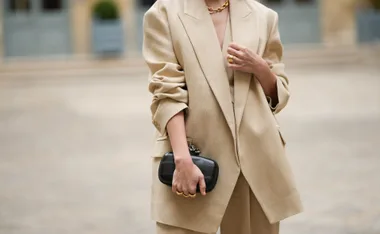 A person wearing a beige blazer with rolled sleeves, holding a black clutch, and accessorized with gold jewelry.