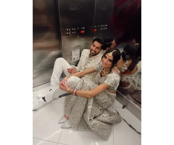 A couple in elegant traditional attire sitting on an elevator floor, smiling and leaning against mirrored walls.