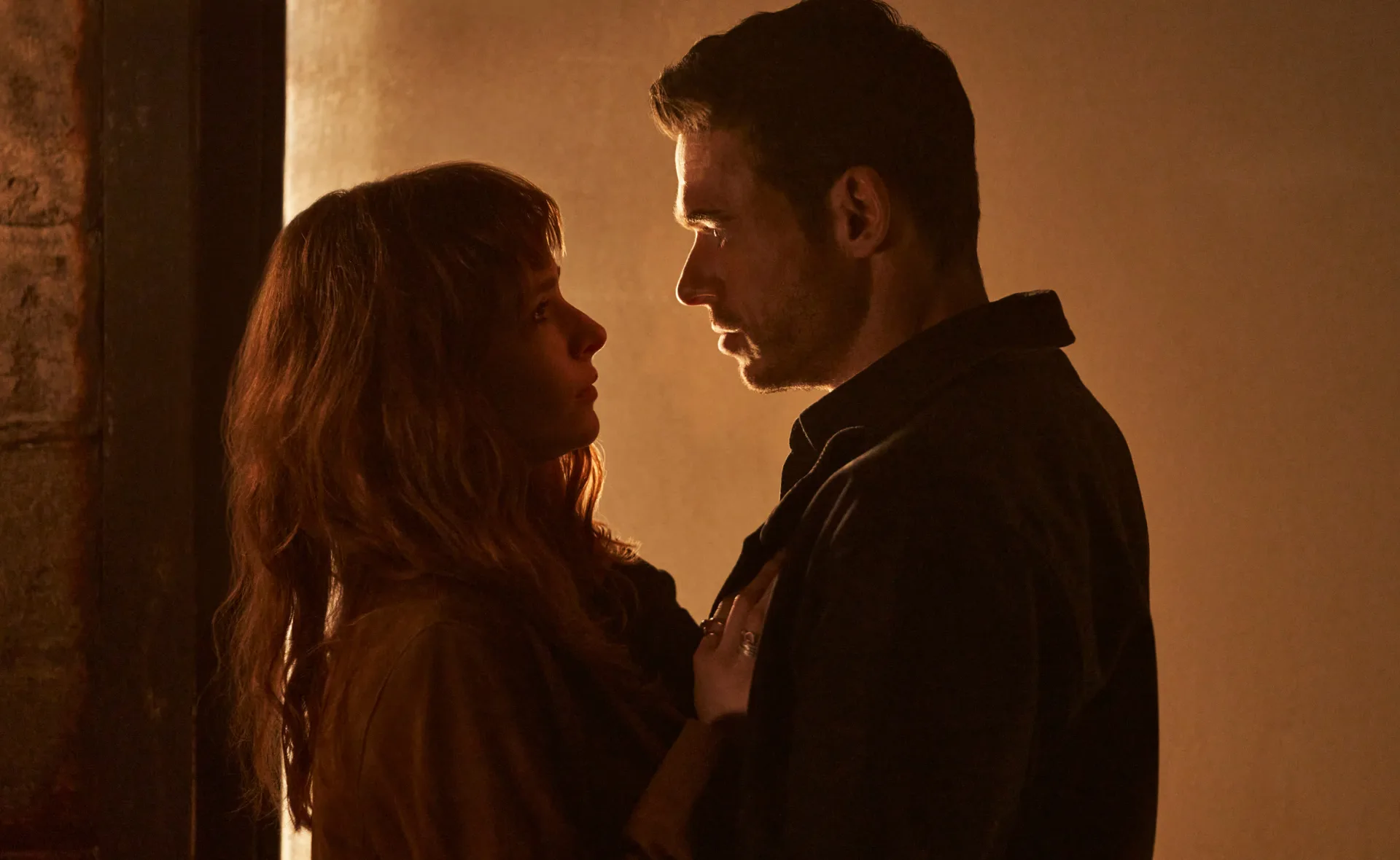 Two people stand closely, gazing into each other's eyes, in a warmly lit setting.