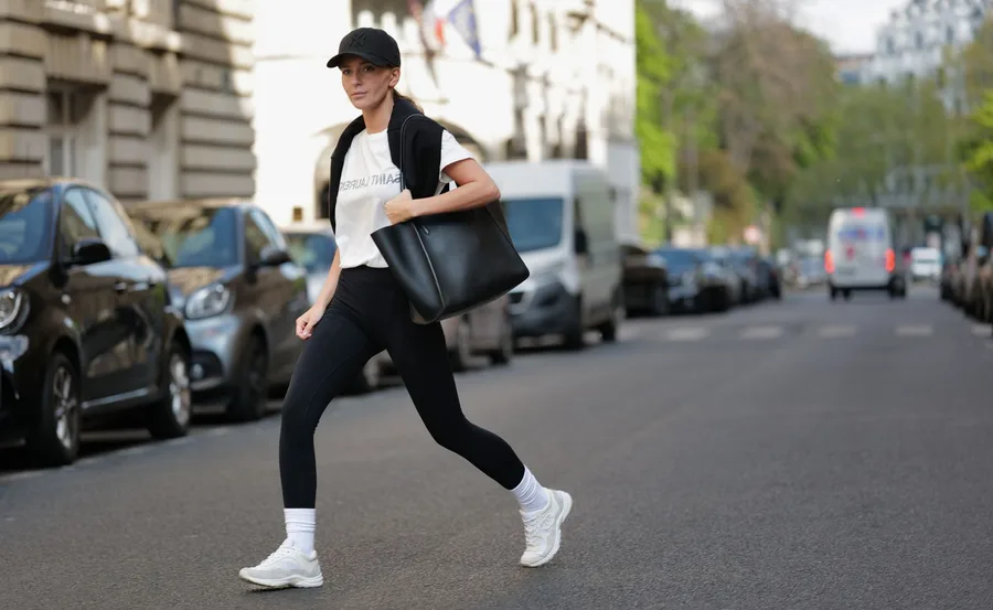 Person crossing a city street wearing leggings, white sneakers, a cap, carrying a large black tote bag.
