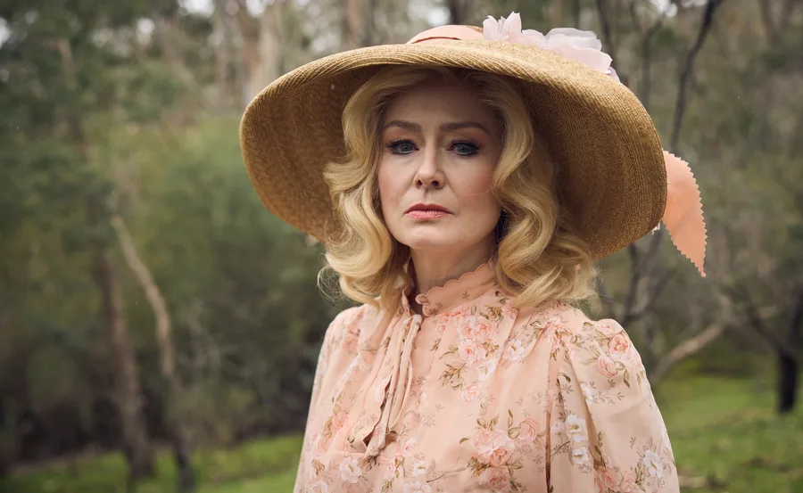 A woman in a floral dress and wide-brimmed hat stands in a forest setting, gazing intently.