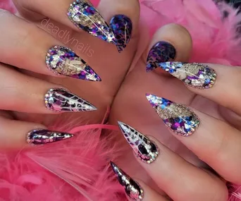 Stiletto nails with vibrant violet, silver glitter, and holographic foil designs on a pink feather background.