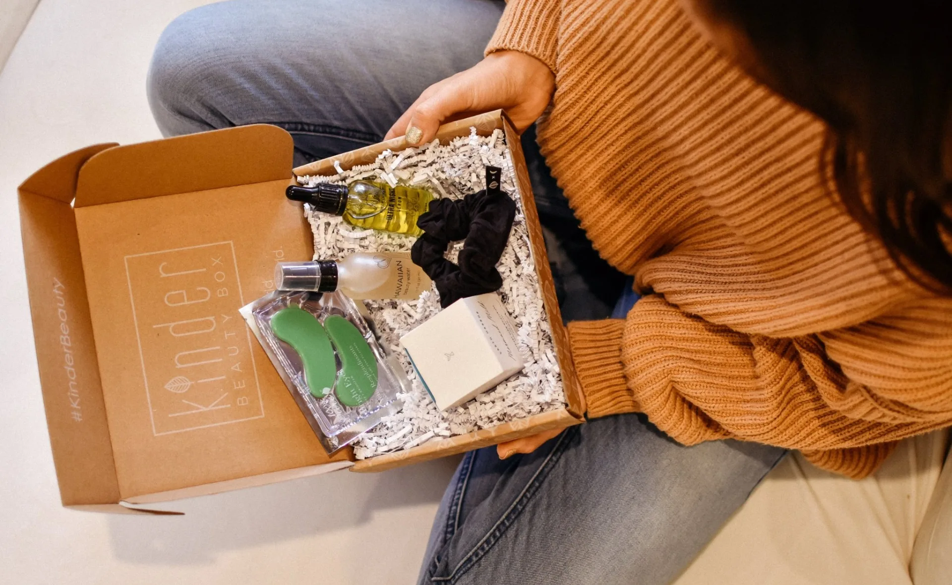 Person holding an open Kinder Beauty Box with skincare products, a scrunchie, and eye masks.