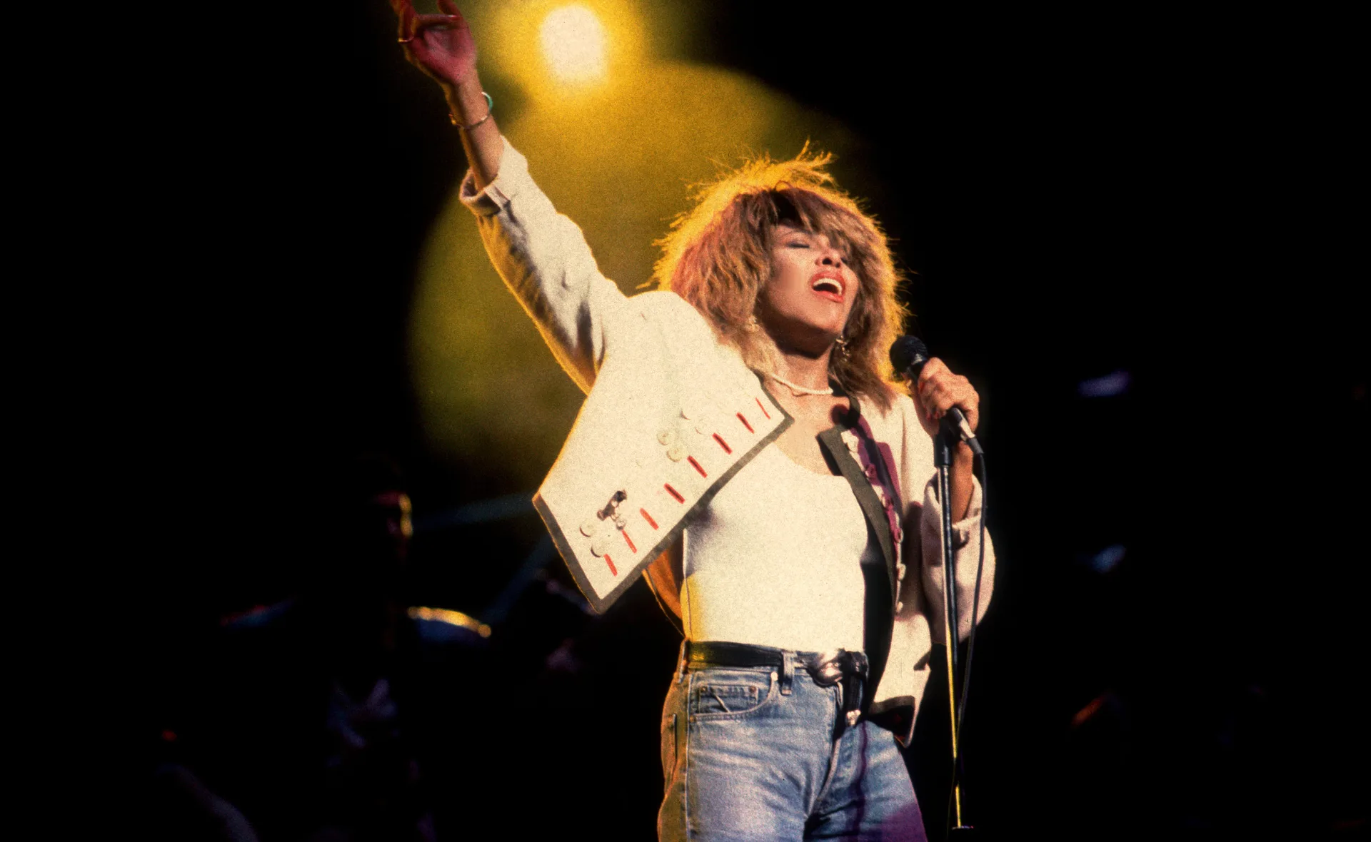 Tina Turner performing energetically on stage in a white outfit under bright stage lights.