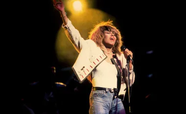 Tina Turner performing energetically on stage in a white outfit under bright stage lights.