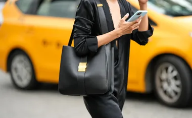 Person in black outfit carrying a black tote bag, using a smartphone, with a yellow car in background.