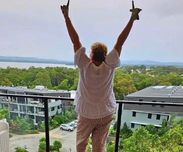 Person on balcony raises arms in celebration, overlooking green landscape and water.