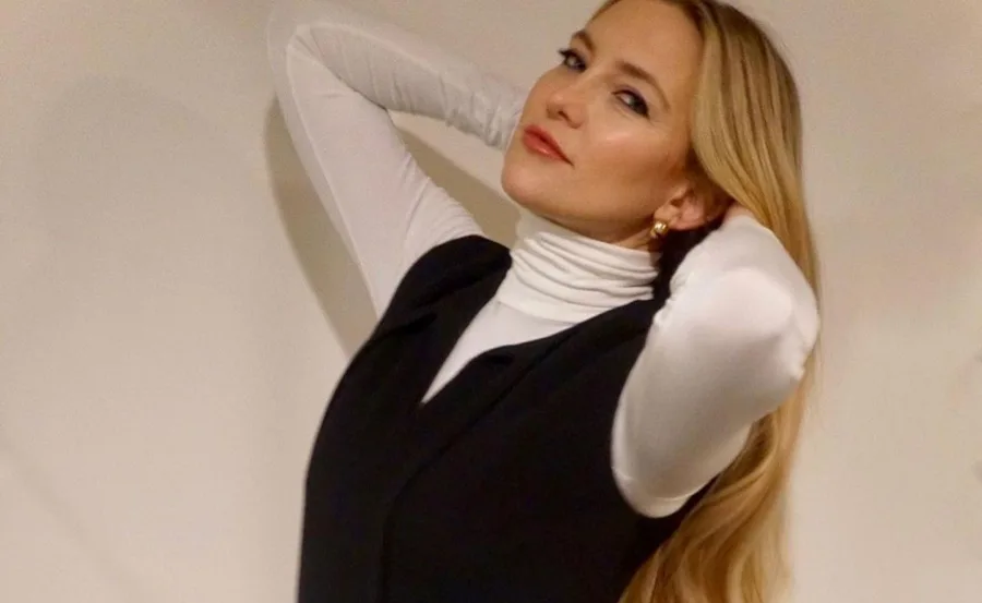 Person with long blonde hair, wearing a white turtleneck and black vest, poses with hands behind head against a plain background.