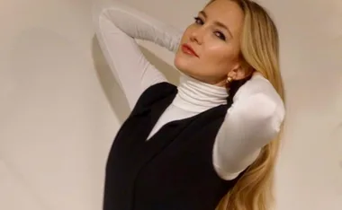 Person with long blonde hair, wearing a white turtleneck and black vest, poses with hands behind head against a plain background.