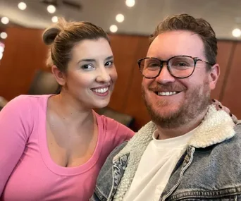 A smiling couple poses for a selfie in a warmly lit room, with him wearing glasses and a denim jacket, and her in a pink top.