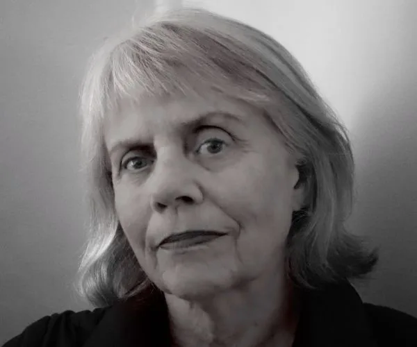 Older woman with short gray hair and a neutral expression, wearing a dark top, against a plain background.