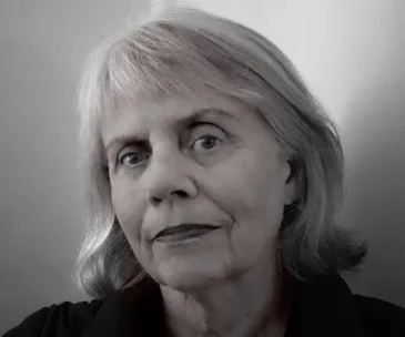 Older woman with short gray hair and a neutral expression, wearing a dark top, against a plain background.