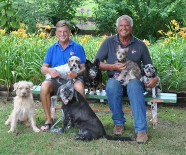 Two men sitting on a bench in a garden, surrounded by six happy dogs.