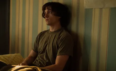 Young man sitting against a striped wall, looking pensive, in a dimly lit room.