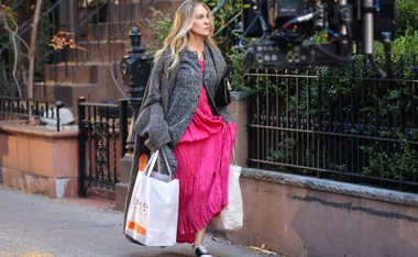 A woman in a pink dress and coat, holding shopping bags, walks on a city street with film equipment nearby.