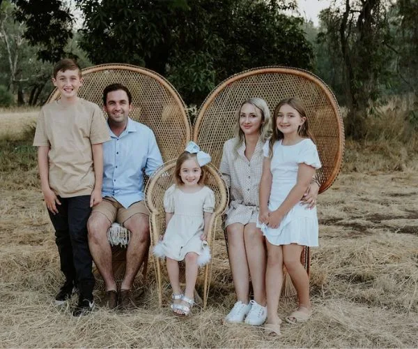 Family of five sitting and standing outdoors on wicker chairs, surrounded by trees and dry grass.