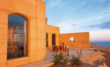 Earth-toned building on Kangaroo Island, tall windows, terrace with plants and ocean view under a blue sky at sunset.