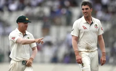 Two Australian cricketers in white uniforms on a field; one points while the other smiles.