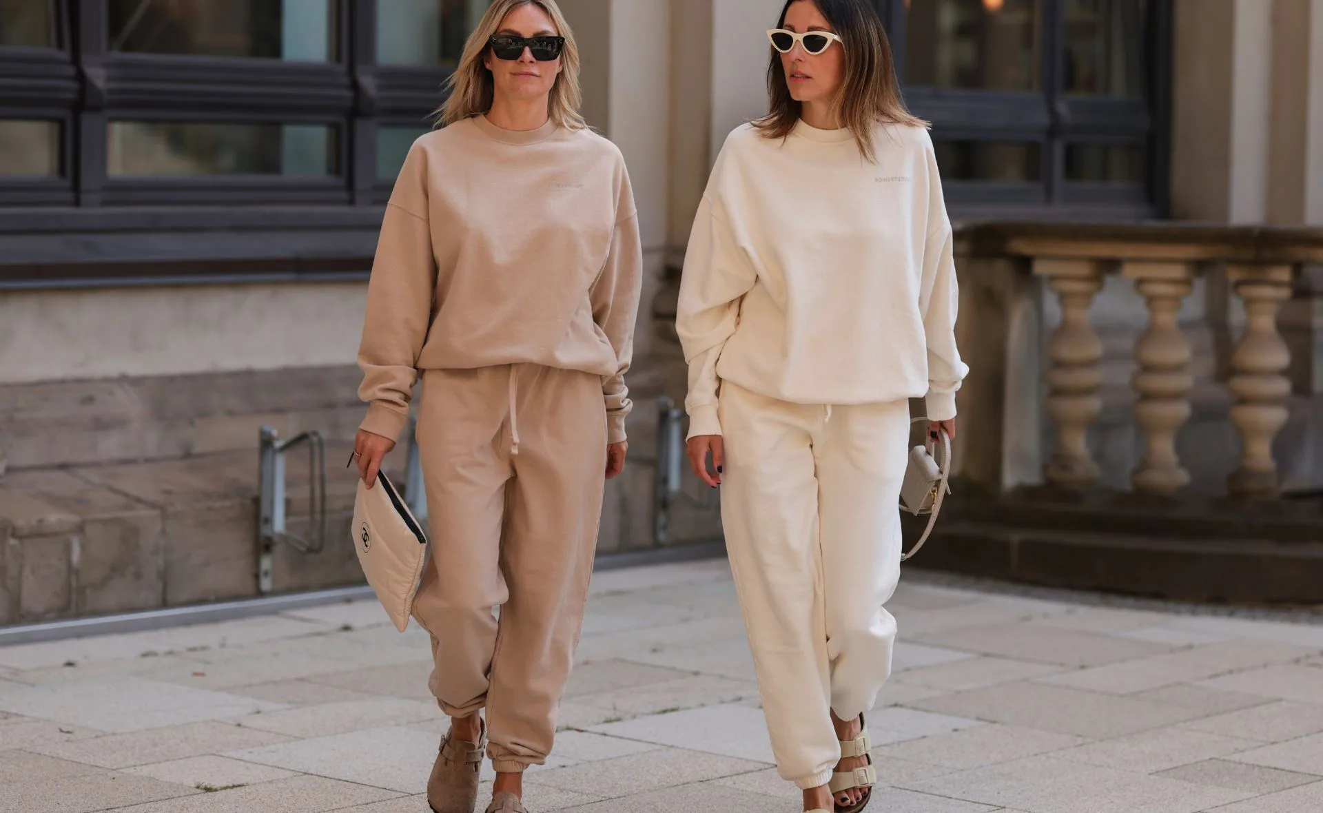 Two women in beige and cream tracksuits walking on a stone pavement, both wearing sunglasses and holding clutches.