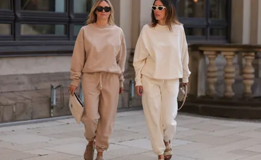 Two women in beige and cream tracksuits walking on a stone pavement, both wearing sunglasses and holding clutches.