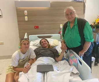 A woman in a hospital bed smiling, holding hands with two visitors, one seated and another standing.