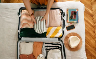 Packing a suitcase with striped and colorful clothing, books, sunglasses, a phone, and a hat on the side.