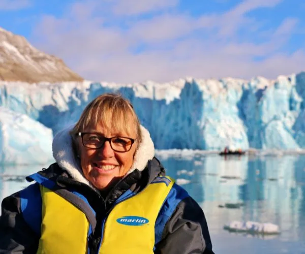 Person in a yellow life jacket smiles in front of a glacier, with a kayak in the background.