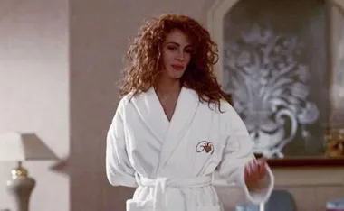Person with curly hair wearing a white dressing gown in an elegant room, reflecting a classic movie scene.