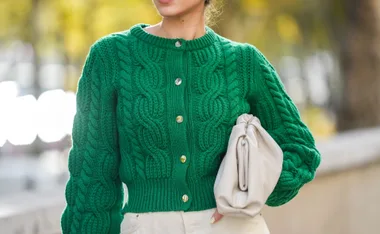Woman in a green cable-knit cardigan holding a white clutch, standing outdoors.