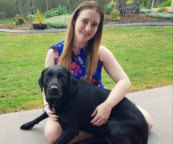 Woman sitting on grass with a black Labrador retriever in her lap, in a garden setting.
