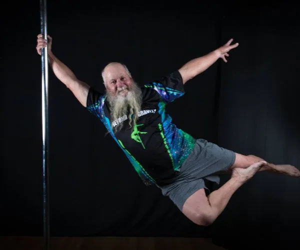 Elderly man with long beard performs a pole dance move, wearing colorful shorts and shirt, against a dark background.