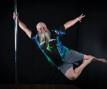 Elderly man with long beard performs a pole dance move, wearing colorful shorts and shirt, against a dark background.