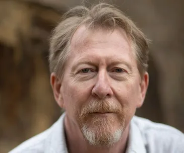 A middle-aged man with short hair and a beard, wearing a light shirt, looking at the camera with a neutral expression.