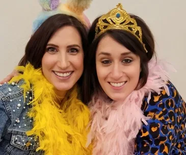 Two smiling women wearing colorful boas and a gold crown, embracing each other at a fun gathering.