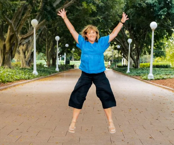 Person joyfully doing a star jump on a tree-lined pathway with lamps.