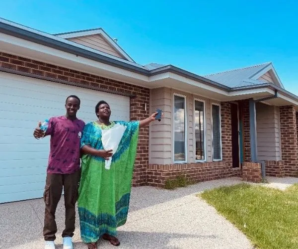 Man and woman standing happily outside a new brick house, holding house keys and paperwork.