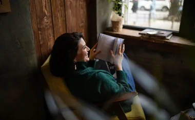 A woman in a green sweater reads a book while seated in a cozy chair by a window.