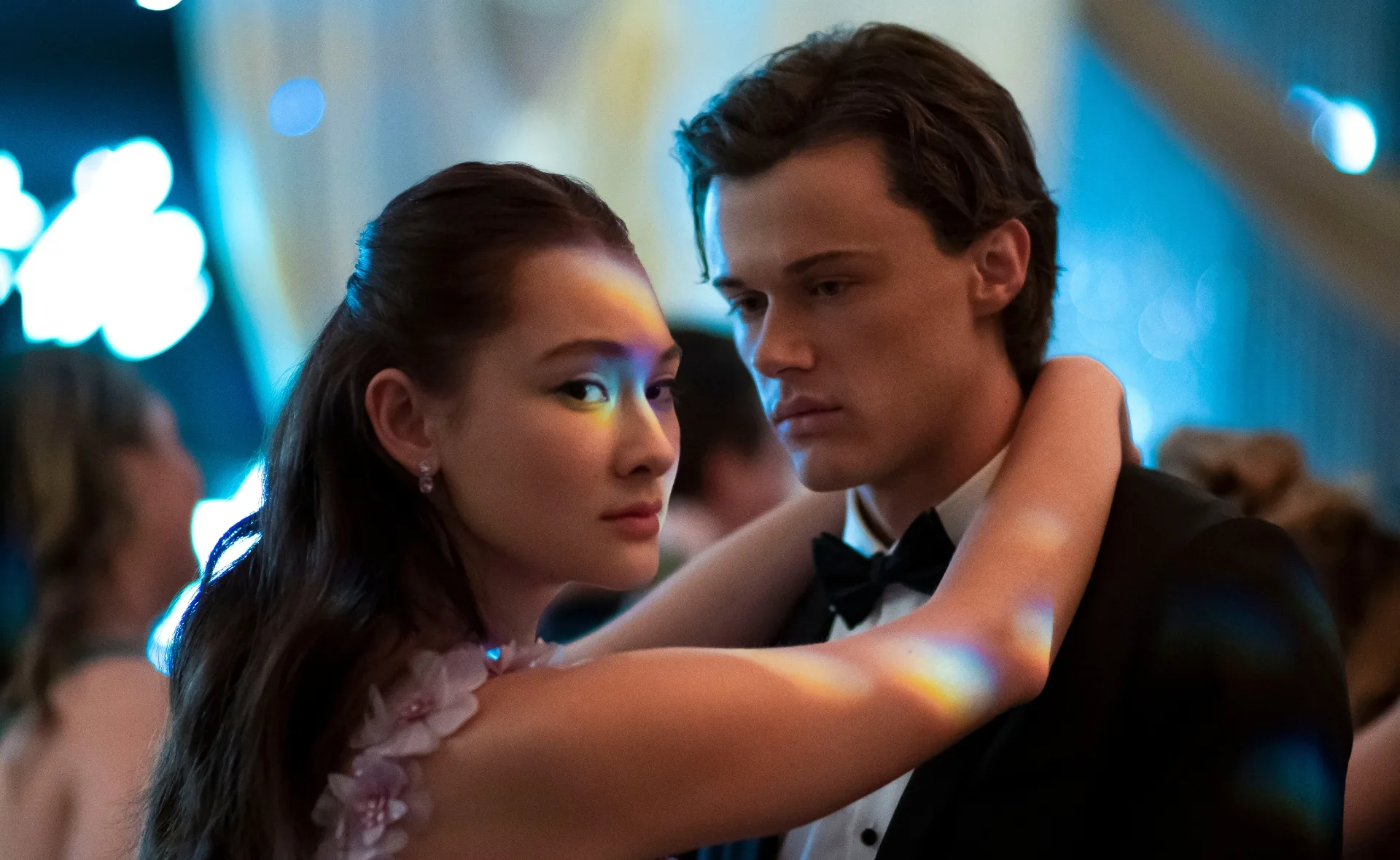 Two young people dancing closely at a formal event, with colorful lighting reflecting on them.