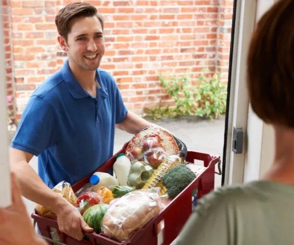 Man delivering groceries in a basket to a woman at the door; includes bread, vegetables, milk, and other items.