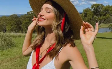 Woman in a sun hat enjoying nature with eyes closed, wearing a red scarf and white top in a grassy field.