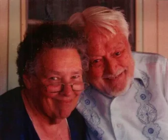Elderly couple smiling, the man has white hair and the woman wears glasses, both appear happy and content.