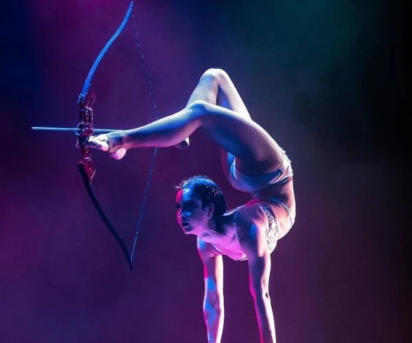 Contortionist performs a backbend, holding a bow with feet, poised to shoot an arrow.
