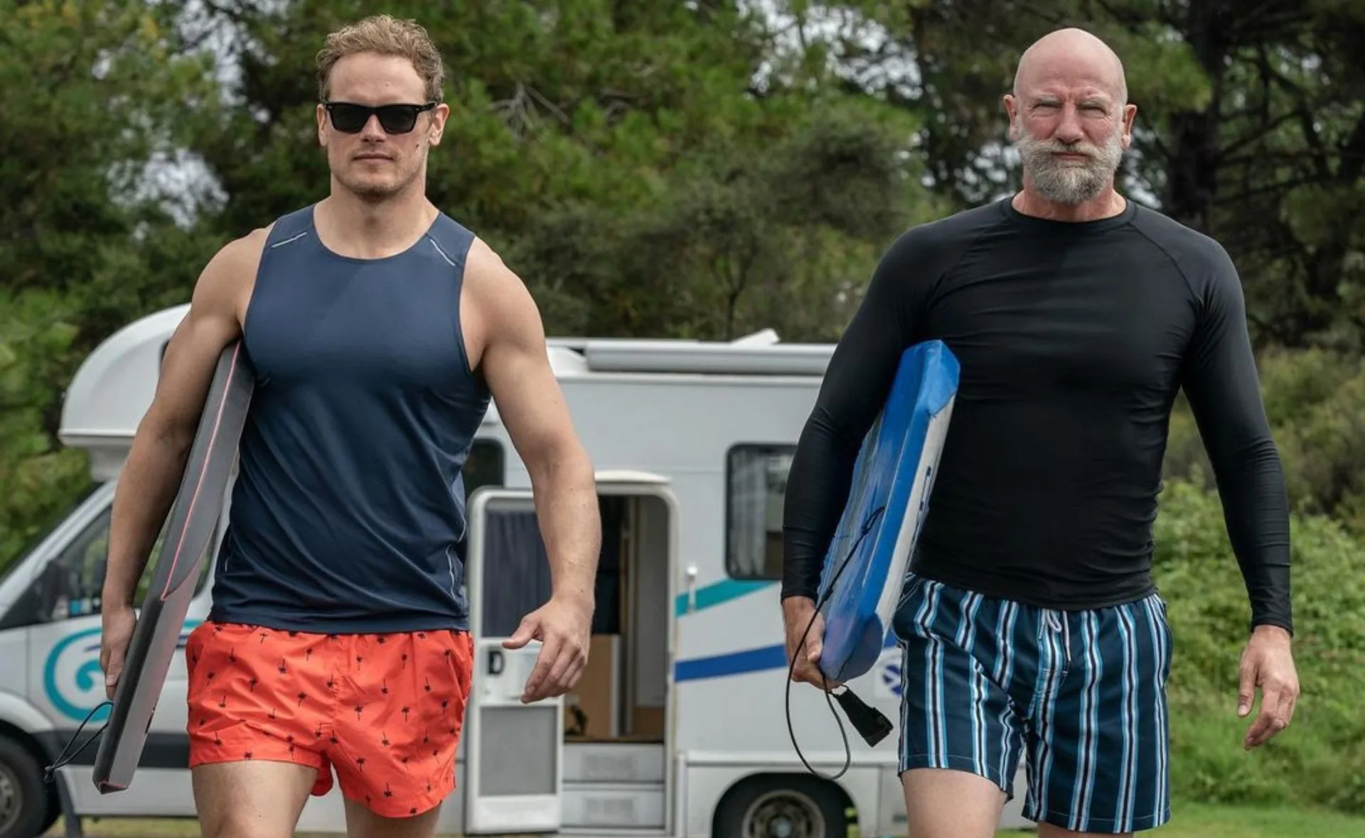 Two men wearing board shorts and tank tops carrying surfboards, walking near a camper van in a leafy area.