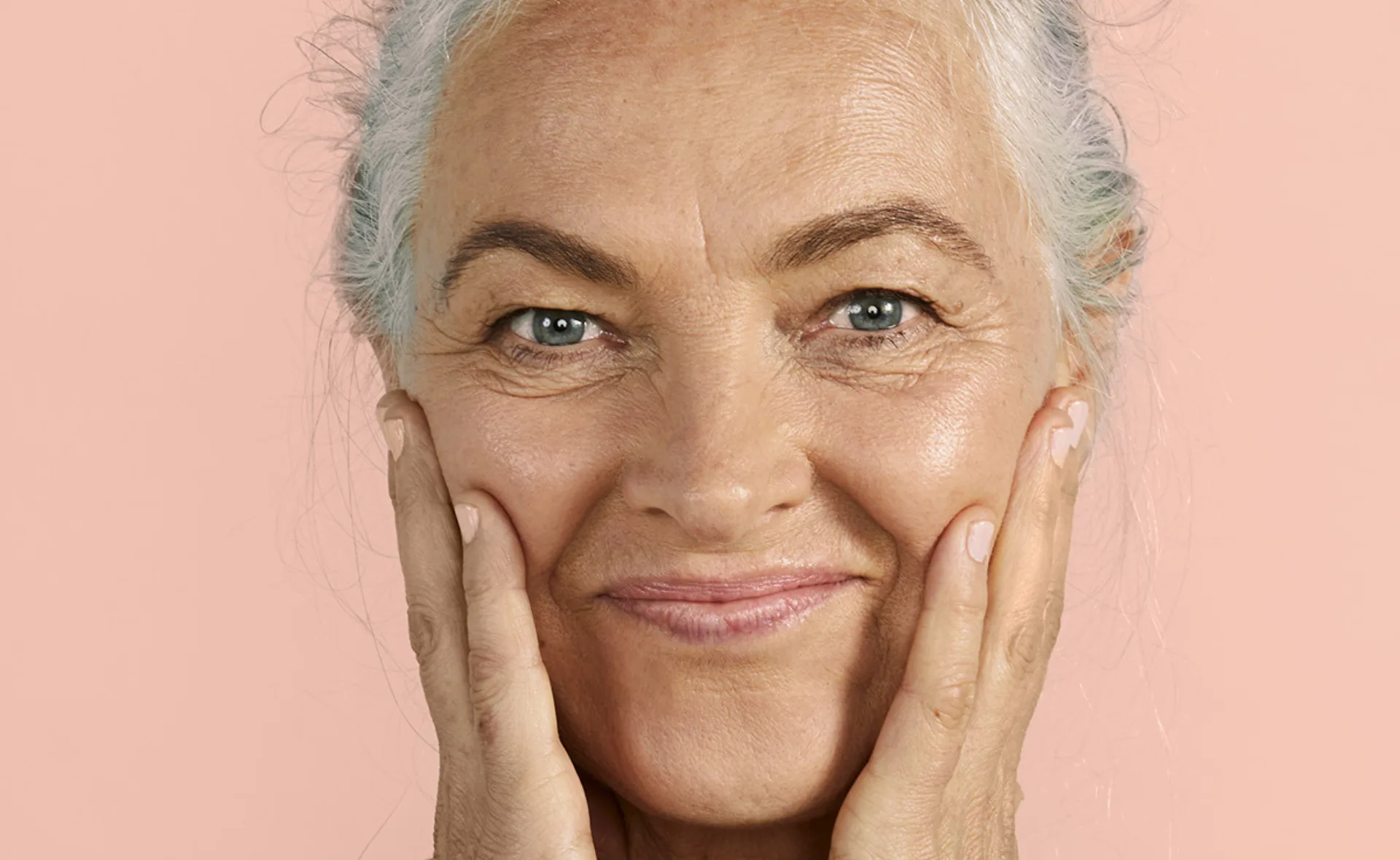"Older woman with gray hair smiling and gently touching her cheeks against a peach background."