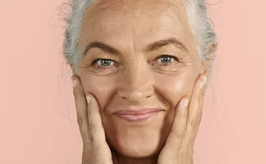 "Older woman with gray hair smiling and gently touching her cheeks against a peach background."