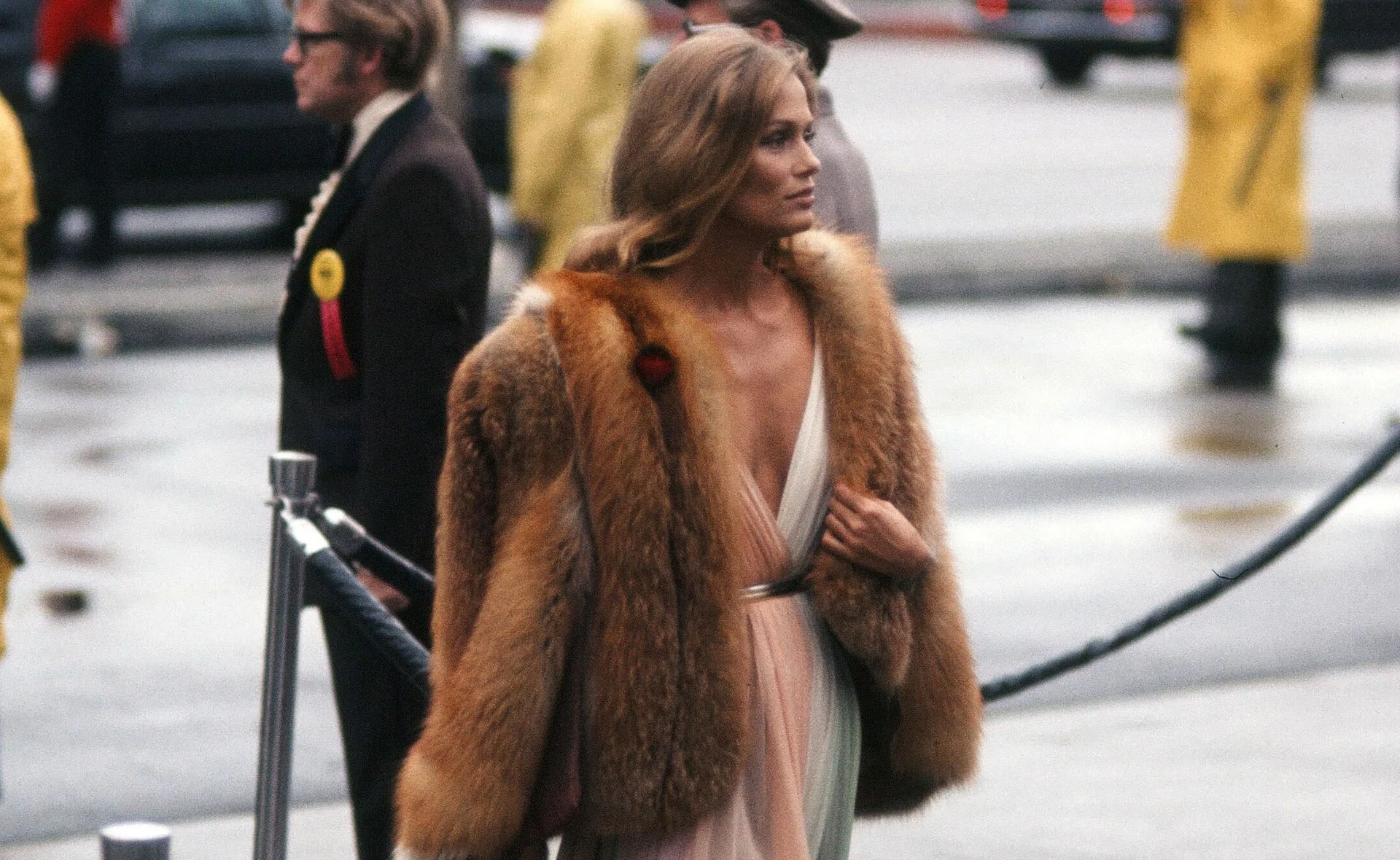 A woman in a fur coat and deep V-neck dress walks past a crowd, with a man in glasses in the background.
