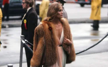 A woman in a fur coat and deep V-neck dress walks past a crowd, with a man in glasses in the background.