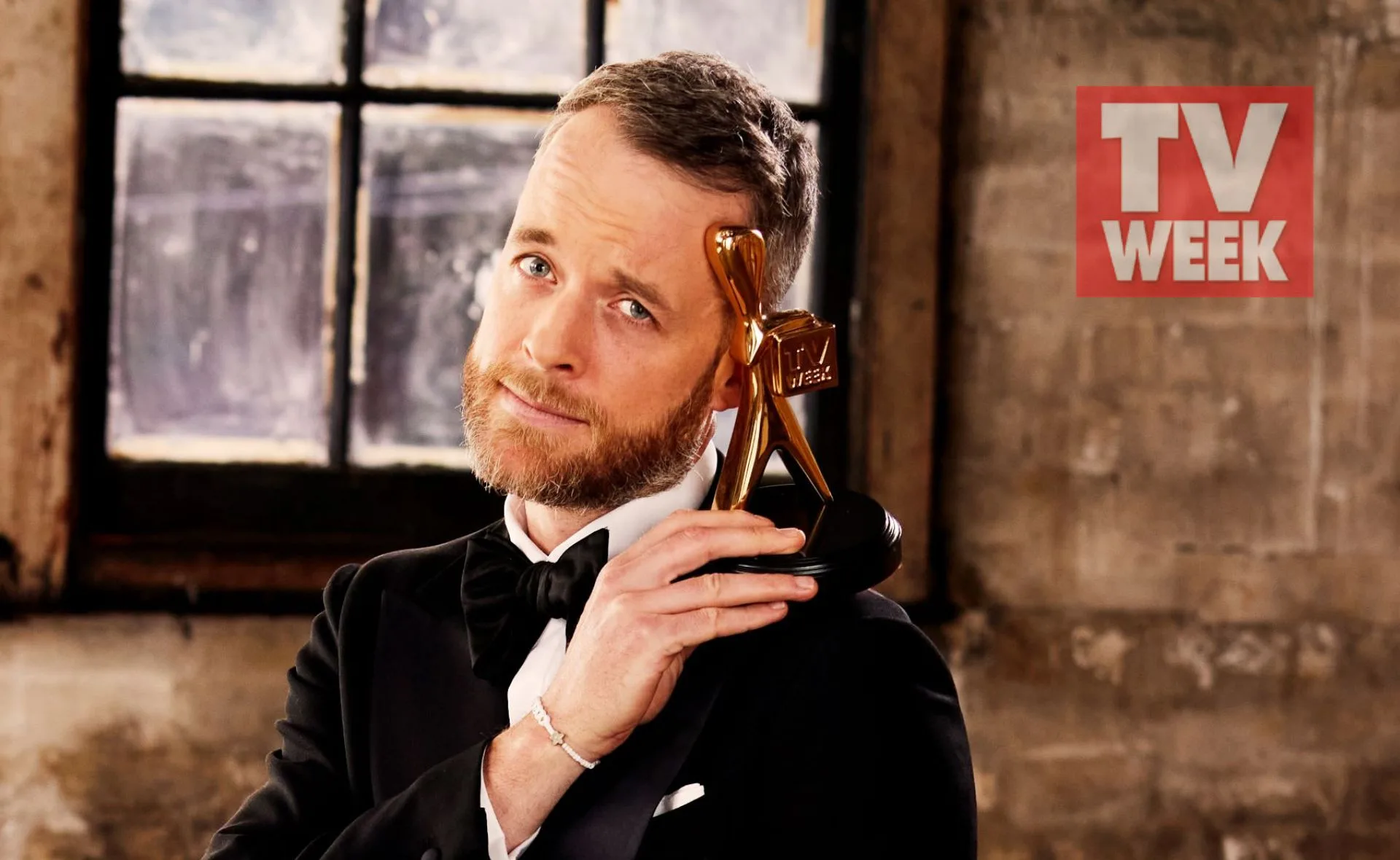 A person in a tuxedo holds a TV Week Logie award, posing against a rustic window background.